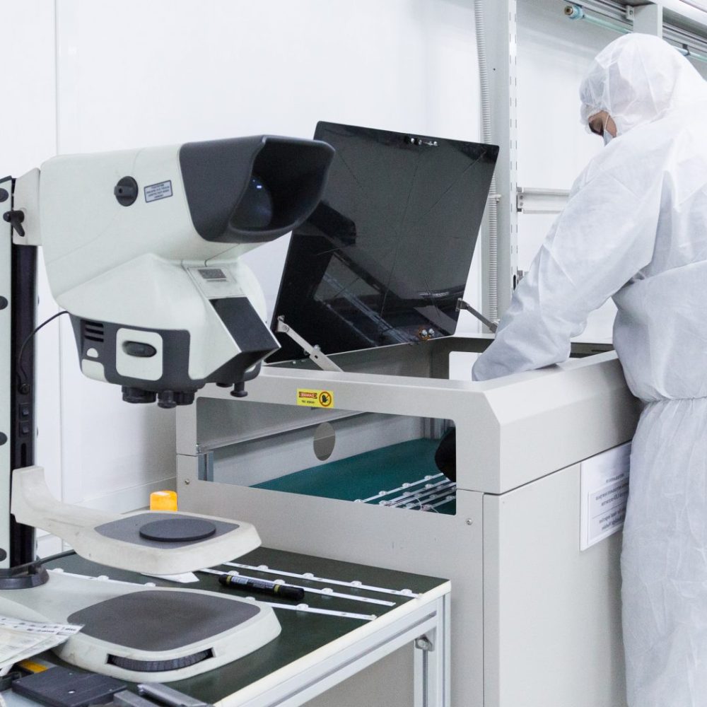 a factory worker in a white lab suits and face masks, working with some modern equipment in a clean white room. an electron microscope is in the foreground.